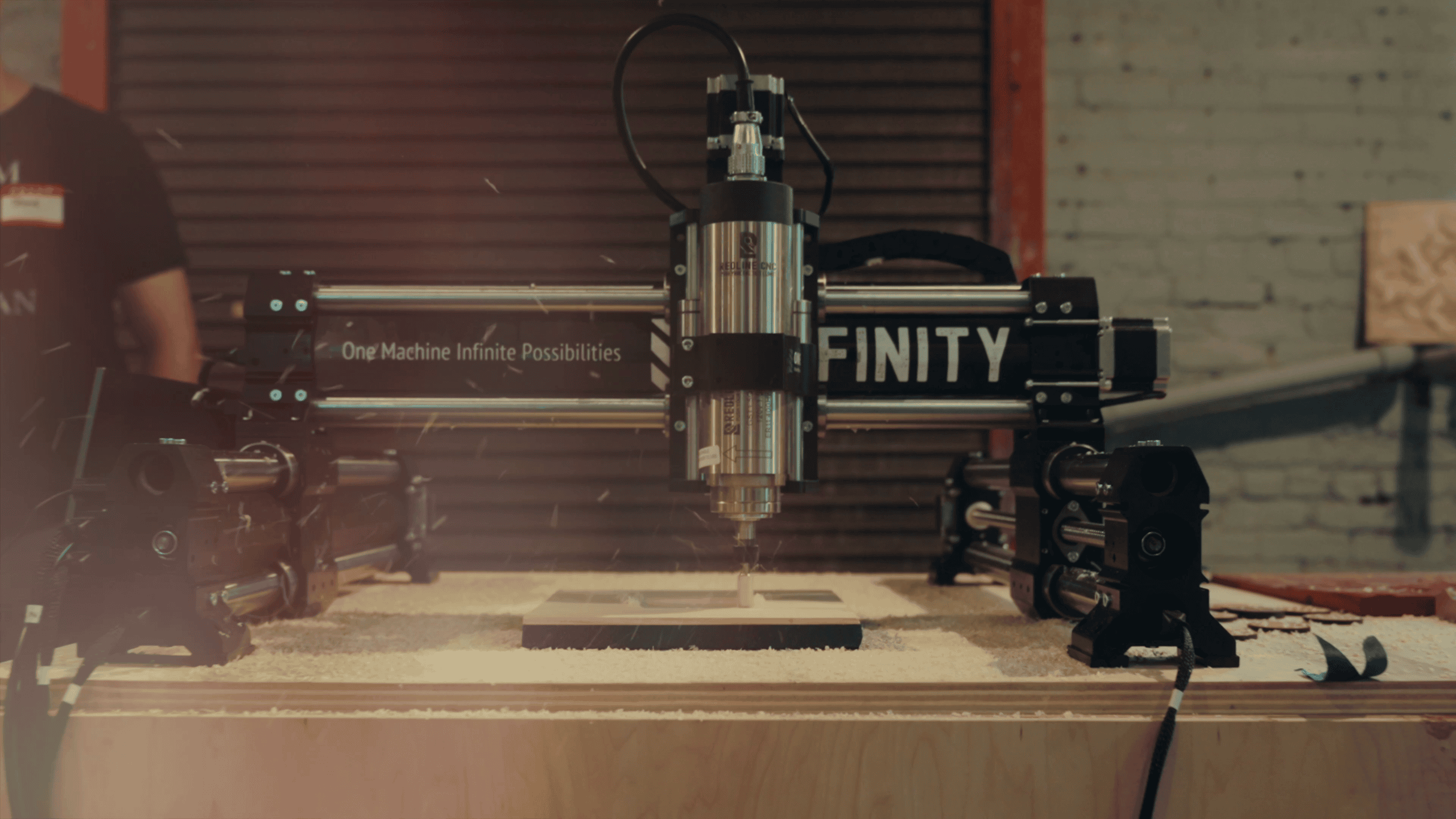 A Onefinity CNC machine carving a piece of wood, sawdust flying, with 'One Machine Infinite Possibilities' printed on the gantry