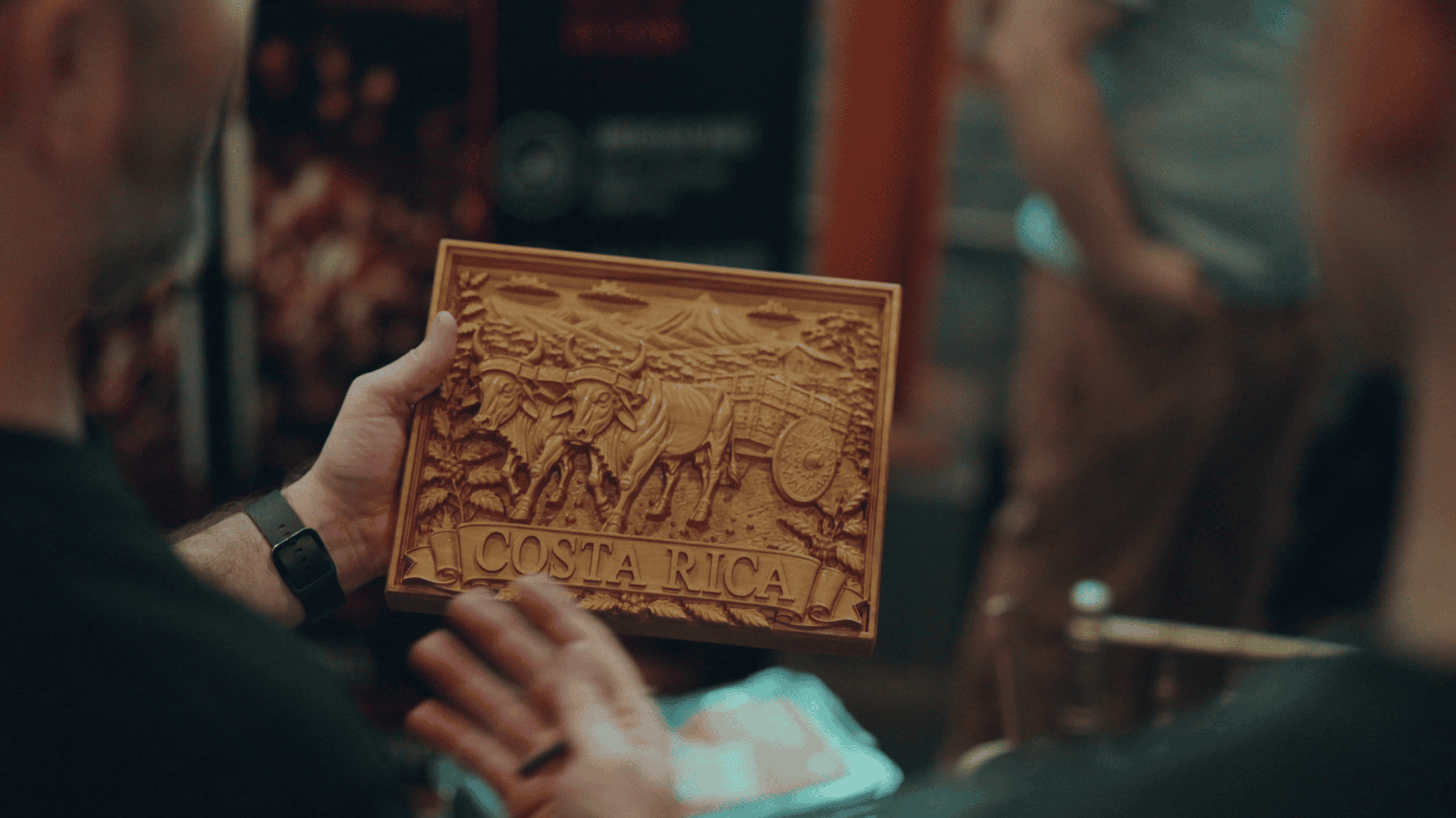 UG Still 11 A detailed wood carving depicting an oxcart and mountains, with 'Costa Rica' carved into a banner at the bottom, held up by an attendee